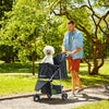 A man is pushing a black 3-wheel foldable pet stroller with mesh windows through a grassy park path.