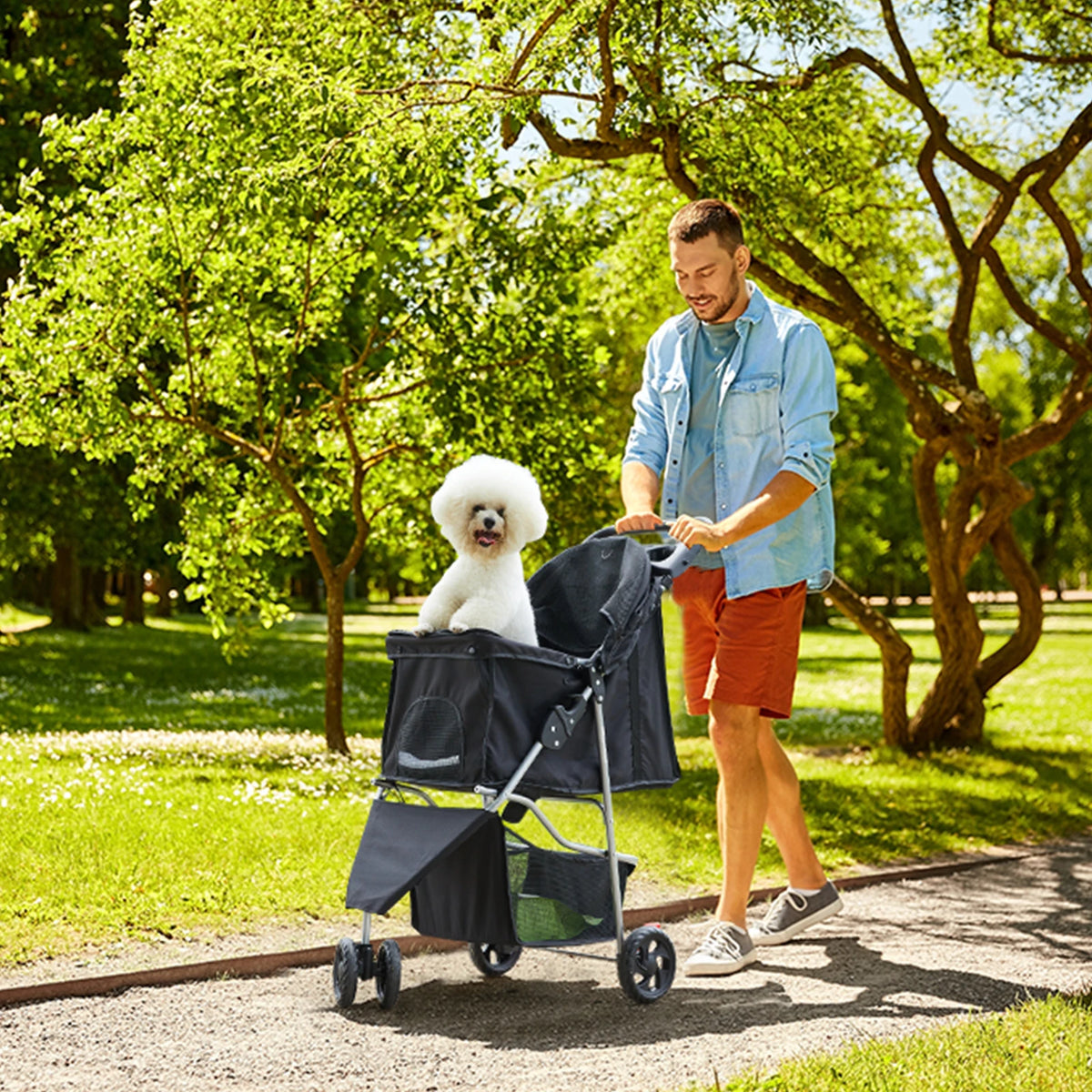 A man is pushing a black 3-wheel foldable pet stroller with mesh windows through a grassy park path.