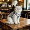 A fluffy grey cat wearing a navy and white striped knitted vest sitting on a wooden table.