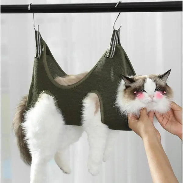 A fluffy white and gray cat is hanging from a green pet grooming hammock for nail clipping.