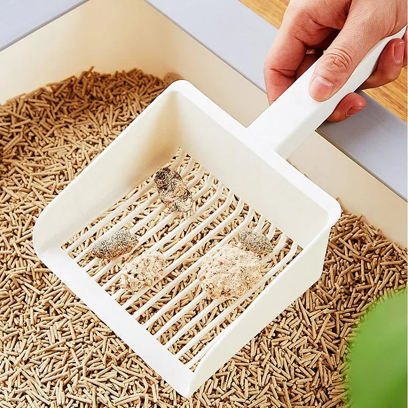 A hand is using an extra-large white cat litter scoop with a hanging hook to scoop litter from a box of wood pellets.