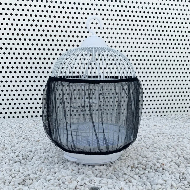 A white mesh bird cage with a black fabric guard is displayed on a light-colored gravel surface.
