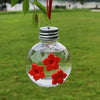 Un distributeur d'eau sphérique en verre clair, rempli d'eau et décoré de fleurs rouges, suspendu à une branche de plante.