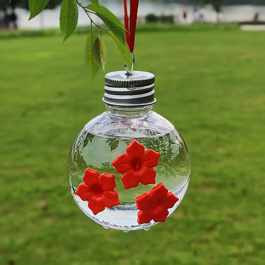 Un distributeur d'eau sphérique en verre clair, rempli d'eau et décoré de fleurs rouges, suspendu à une branche de plante.