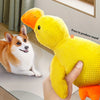 A yellow duck plush toy is being held by a hand, with a dog sitting nearby.