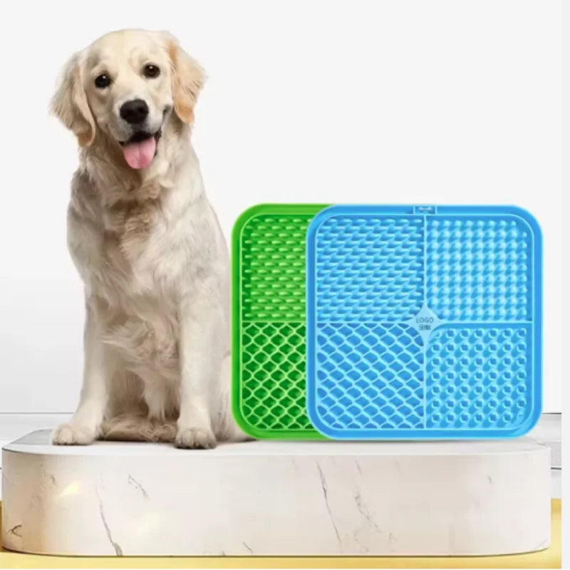 A light-colored dog is sitting next to a green and blue silicone lick mat for training.