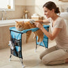 A woman is using a foldable pet grooming hammock to trim the nails of an orange cat in a kitchen setting.