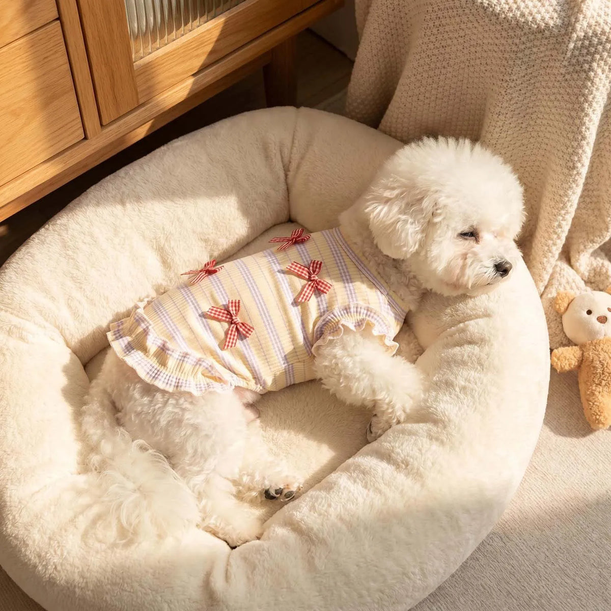 A fluffy white dog wearing a yellow and white patterned mesh bow suit resting in a cream-colored pet bed.