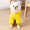 A fluffy white dog wearing a bright yellow fleece jumpsuit and hood, sitting on a light wooden floor.