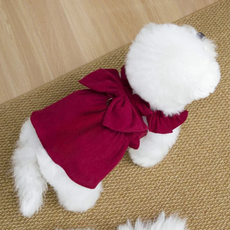 A white fluffy dog toy wearing a deep red corduroy pet dress with a bow and ruffle skirt.
