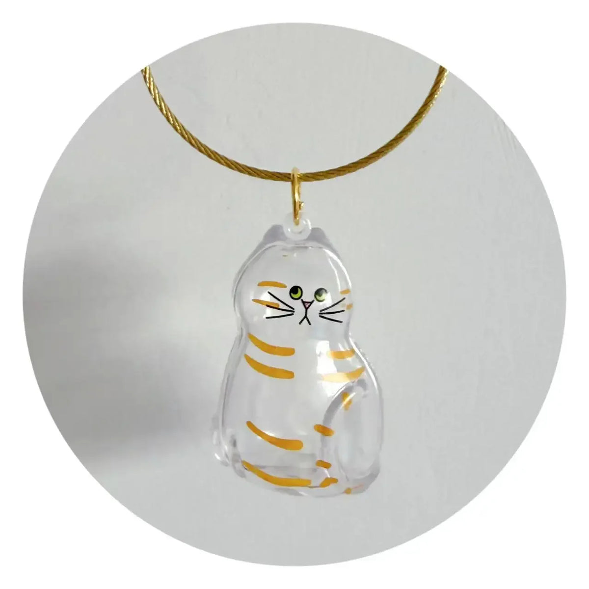 Un charm en verre clair en forme de chat rayé suspendu à une corde dorée.