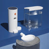 A white automatic pet drinking water dispenser is shown next to a clear container and a gray feeding dish on a blue surface.