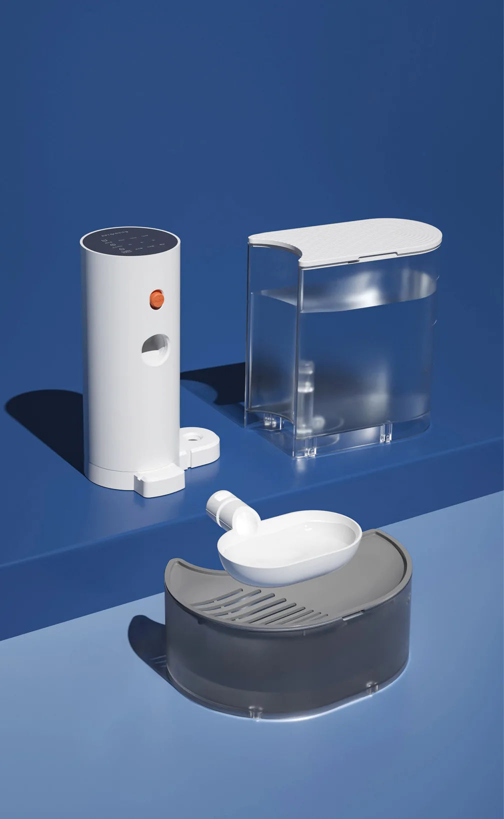 A white automatic pet drinking water dispenser is shown next to a clear container and a gray feeding dish on a blue surface.