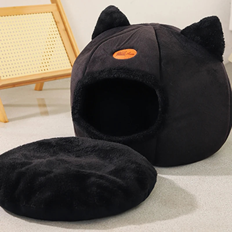 A black plush cat cave bed with a washable cushion and ear flaps is shown on a light surface.