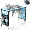 A gray cat is relaxing in a blue pet grooming hammock suspended from a stand, with water being sprayed onto the cat.
