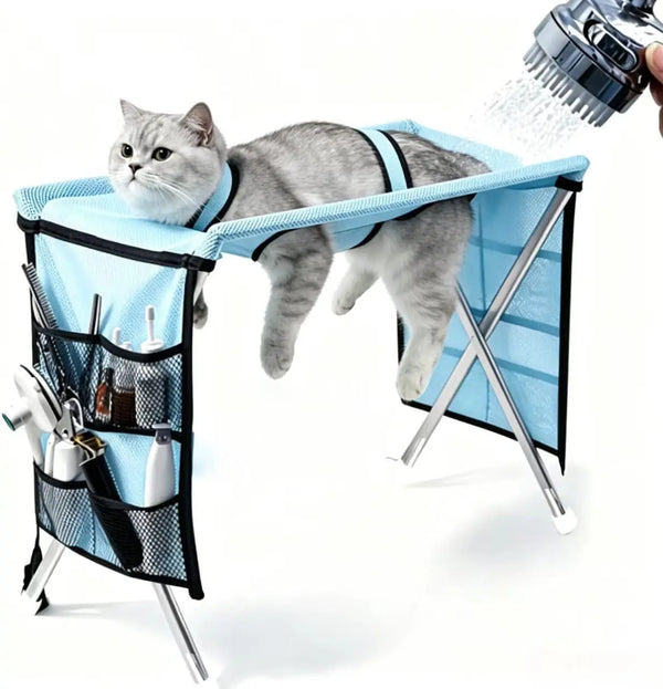 A gray cat is relaxing in a blue pet grooming hammock suspended from a stand, with water being sprayed onto the cat.