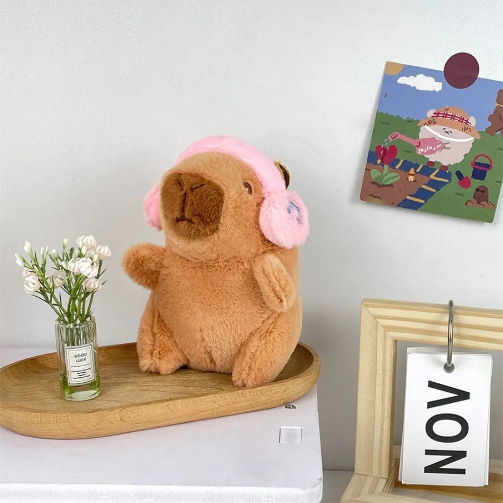 A fluffy brown capybara plush doll wearing a pink hoodie and wings sitting next to a framed picture.