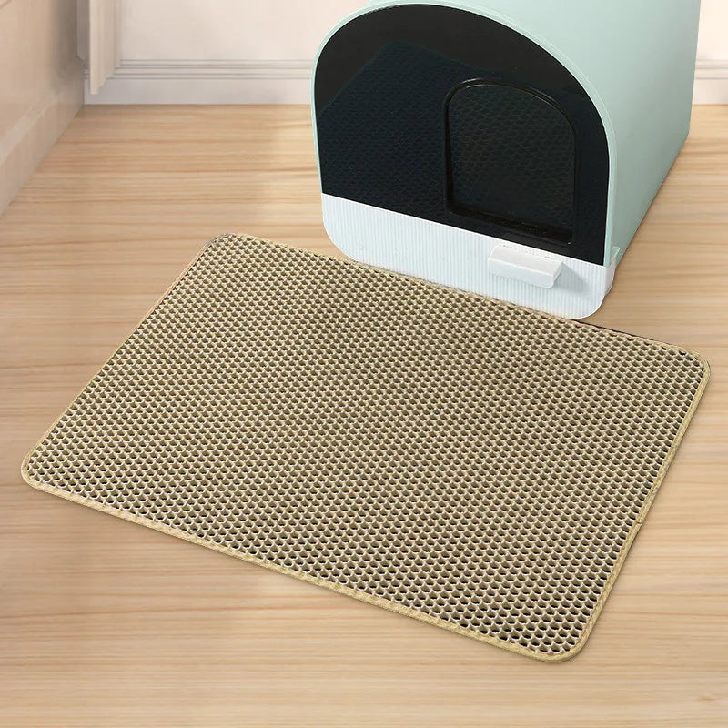 A beige double-layer cat litter mat is placed on a wooden floor next to a white and black pet grooming device.