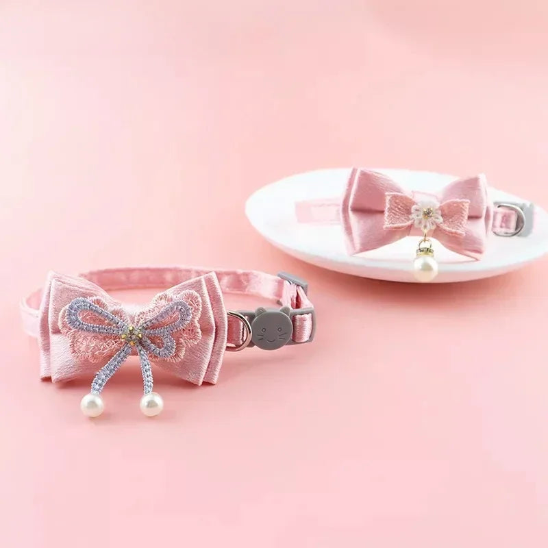 Pinkes Spitzen-Halsband für Haustiere mit Schleife, Hängerkörnen und einer Glocke auf einer weißen Platte.