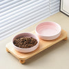 A pink ceramic bowl filled with dark kibble sits on a wooden stand, next to a window blind.