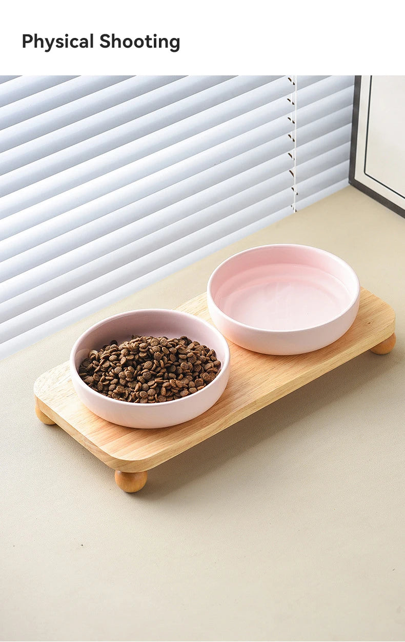 A pink ceramic bowl filled with dark kibble sits on a wooden stand, next to a window blind.