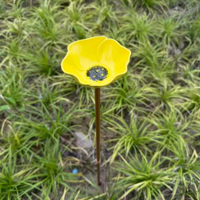 A yellow flower with a black center is displayed on a thin metal stem against a background of green grass.