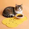 A tabby cat is looking at a pink bowl filled with dry pet food on a yellow mat.