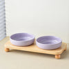 A light purple ceramic bowl is placed on a natural wood shelf, used as a stand for a pet bowl.