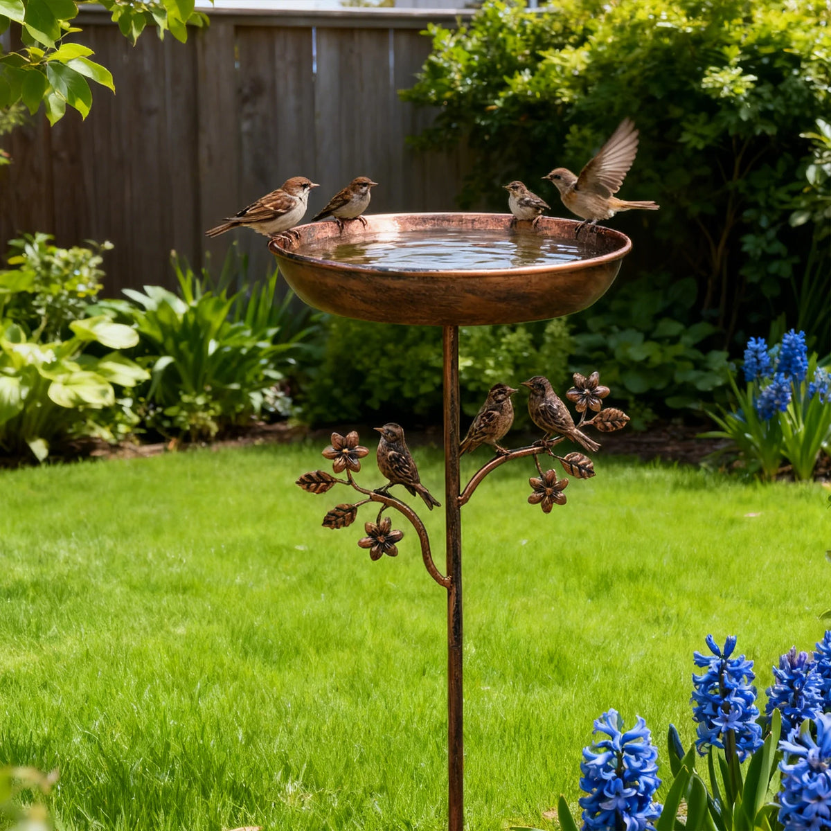 Kupfer-Vogelbad mit kleinen Vögeln auf dem Rand in einer üppigen Gartenumgebung.