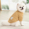 A white dog wearing a knitted tan sweater sitting on a light surface.