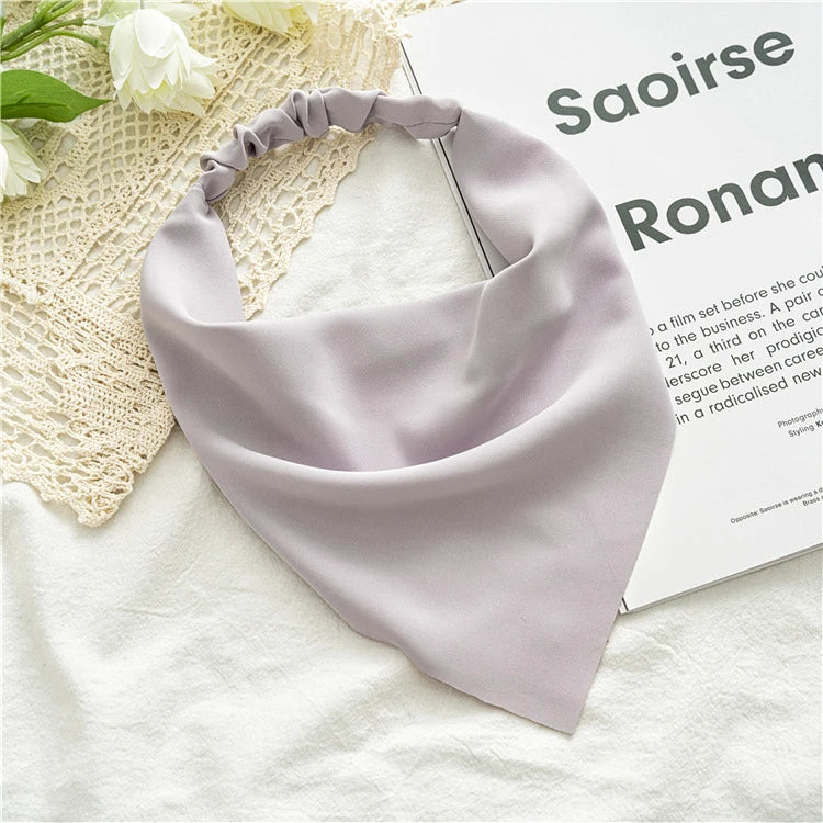 A soft lilac bohemian paisley triangle head scarf is displayed on a white fabric next to a book.