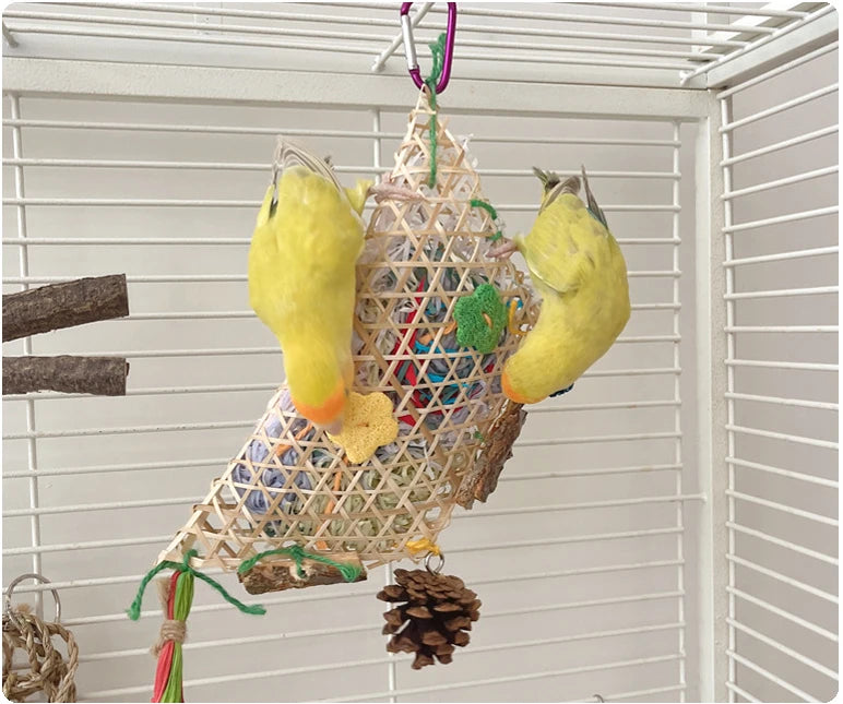A yellow parrot chewing toy with a bamboo frame and colorful shredded paper hanging in a white grid