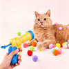 Eine tabbyartige Katze liegt, während eine Hand eine blaue Spielzeugwaffe und mehrere bunte Plüschbälle hält.