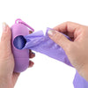 A hand is holding a purple capsule-shaped dog poop bag dispenser with a carabiner clip and a crumpled purple plastic bag.
