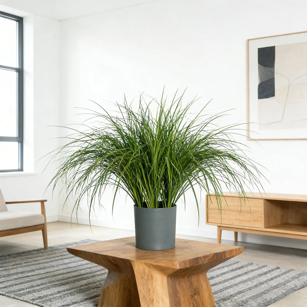 A potted artificial reed grass bouquet in a gray planter sits on a wooden table in a bright room.