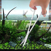 A hand holds a stainless steel aquarium cleaning tool set above a glass tank filled with aquatic plants and debris.