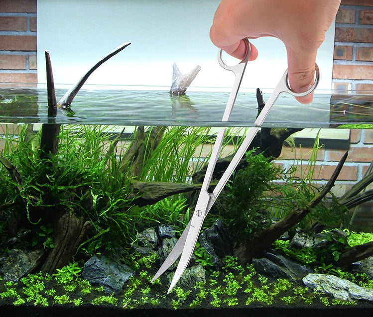 A hand holds a stainless steel aquarium cleaning tool set above a glass tank filled with aquatic plants and debris.