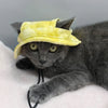 Un gato gris vistiendo un sombrero de sol amarillo ajustable con orificio para las orejas, tumbado sobre pelaje blanco.