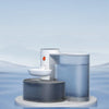 A modern, silver automatic pet drinking water dispenser is shown floating on a blue surface against a pale sky background.