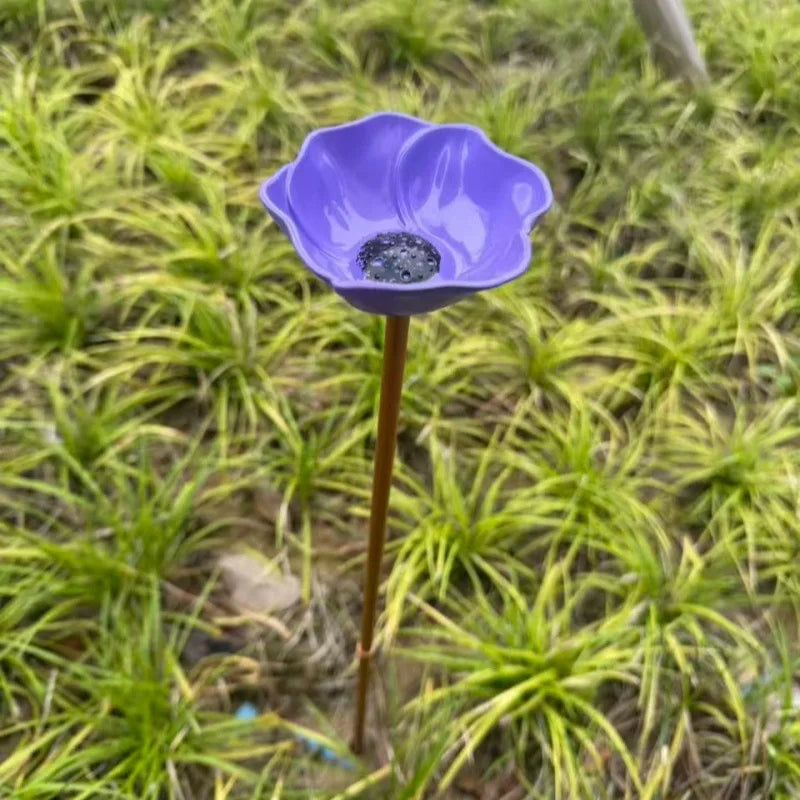 A purple stainless steel bird feeder with a small seed bowl is planted in green grass.