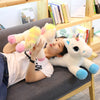 A woman is cuddling a white unicorn plush toy with pink accents on a gray sofa.