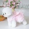 Un perro blanco esponjoso vistiendo un vestido de mascota floral rosa, sentado sobre una superficie blanca.