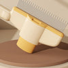 A close-up view of a yellow and white self-cleaning pet brush with massage pins resting on a wooden surface.