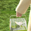 A hand is holding a lime green portable bird carrier bag with a wire mesh cage on a grassy lawn.