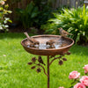 Braune Metallvogelbad mit Wasser, ein Vogel trinkt in einem üppigen grünen Garten.