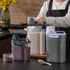 A clear airtight pet food storage container with a measuring cup and a separate container of dry food is shown on a dark surf