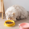 A fluffy gray cat is resting on a wooden surface near a yellow silicone pet feeding bowl and plate.