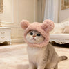 A fluffy, light pink bear cap pet head cover is worn by a cat sitting on a cream rug.