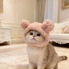 A fluffy, light pink bear cap pet head cover is worn by a cat sitting on a cream rug.