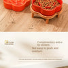 An orange ceramic slow feeder cat bowl with a wooden stand, showing grain being fed into the bowl.